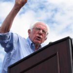 Man speaking at a podium under cloudy sky.