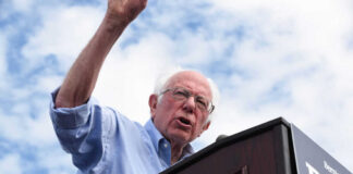 Man speaking at a podium under cloudy sky.