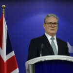 Trump Forces Starmer’s Shock Reversal Man in suit at a podium, British flag behind.
