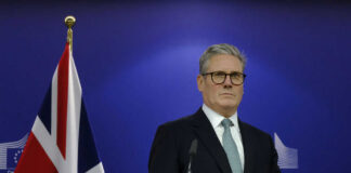 Man in suit at a podium, British flag behind.