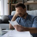 Man sitting in a home office looking stressed while reviewing paperwork