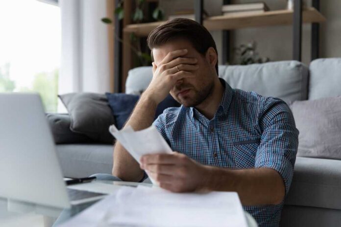 Man sitting in a home office looking stressed while reviewing paperwork