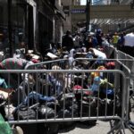 A crowded street with individuals sitting behind barriers and bags, highlighting urban homelessness