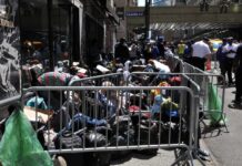 A crowded street with individuals sitting behind barriers and bags, highlighting urban homelessness