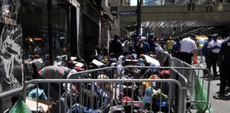 A crowded street with individuals sitting behind barriers and bags, highlighting urban homelessness