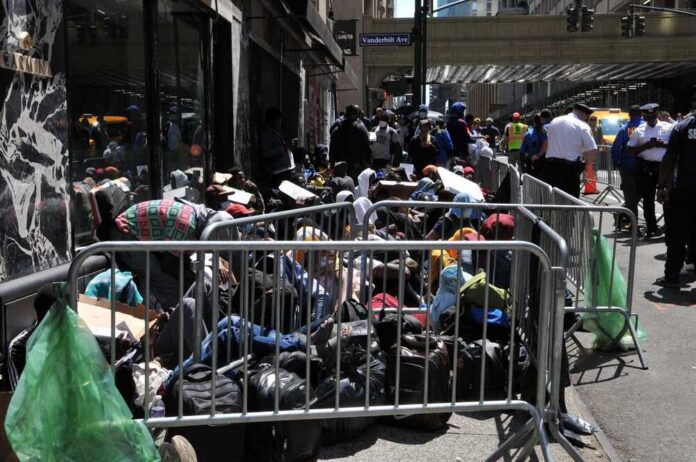A crowded street with individuals sitting behind barriers and bags, highlighting urban homelessness