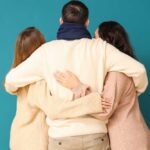 Progressive Icon Normalizes Polyamory—Traditionalists Furious Three people embracing each other from behind against a teal background