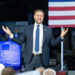 A man in a suit passionately delivering a speech in front of an audience with an American flag in the background