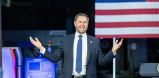 A man in a suit passionately delivering a speech in front of an audience with an American flag in the background