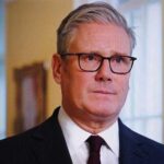 A man in formal attire with glasses, looking serious in an indoor setting