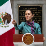 Person speaking at podium beside Mexican flag