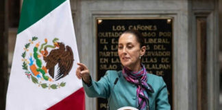 Person speaking at podium beside Mexican flag