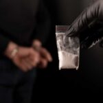 A gloved hand holding a bag of white powder in front of a person in handcuffs