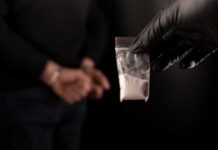 A gloved hand holding a bag of white powder in front of a person in handcuffs