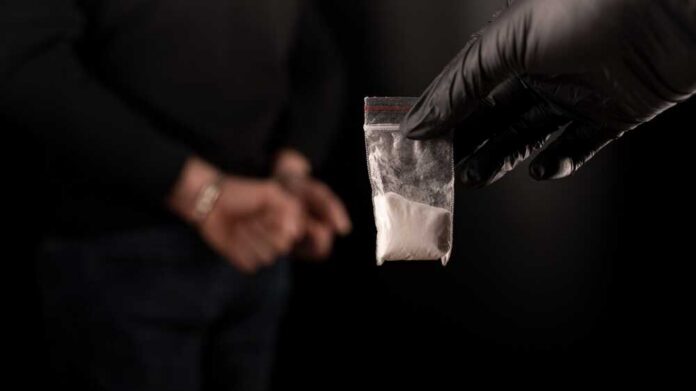 A gloved hand holding a bag of white powder in front of a person in handcuffs