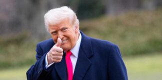 A man in a dark coat and pink tie giving a thumbs up outdoors