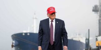 A man in a suit wearing a red cap stands near a ship at a harbor