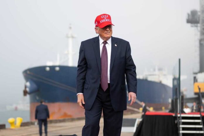 A man in a suit wearing a red cap stands near a ship at a harbor