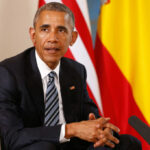 Man in a dark suit seated with flags in the background