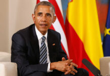 Man in a dark suit seated with flags in the background