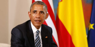 Man in a dark suit seated with flags in the background