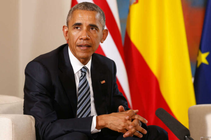 Man in a dark suit seated with flags in the background