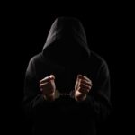 A hooded figure with hands in handcuffs against a dark background