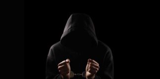 A hooded figure with hands in handcuffs against a dark background