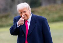 A man in a dark coat and pink tie giving a thumbs up outdoors