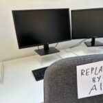 An office desk with two computer monitors and a chair labeled 'REPLACED BY AI'