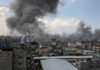 Children Killed; Israel Cites ‘Terrorist’ — Evidence? Cityscape with smoke rising, buildings, and a mosque.