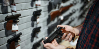 Person holding a handgun in a gun shop with various firearms displayed on the wall