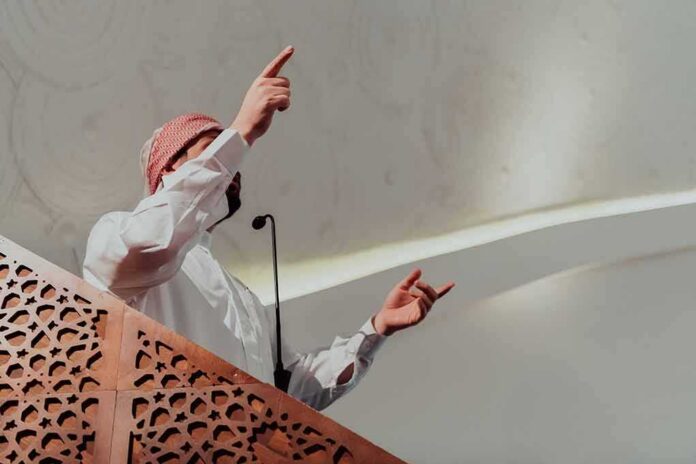 Speaker in traditional attire gesturing from a carved wooden podium