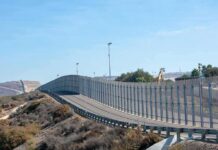 The Border’s New Weapon Moves At Light Speed Tall metal border fence curving through arid landscape
