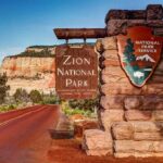 Zion National Park entrance sign beside red road and sandstone cliffs