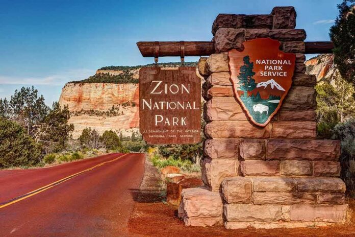 Zion National Park entrance sign beside red road and sandstone cliffs
