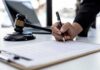 Hand signing legal document beside gavel and laptop
