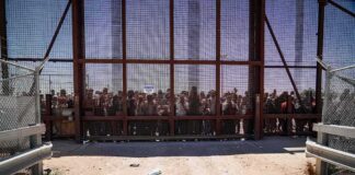 Crowd stands behind tall metal fence at border crossing