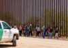Group of people walking along a tall border fence beside a patrol vehicle