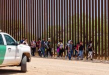 DHS Warning Collides With Growing Caravan Group of people walking along a tall border fence beside a patrol vehicle