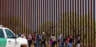 Group of people walking along a tall border fence beside a patrol vehicle
