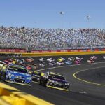 Shock and Sorrow: NASCAR’s Silent Tribute Stock cars racing on a banked track with grandstands in background