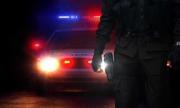 shutterstock_697542187 Police officer stands before patrol car with flashing lights at night
