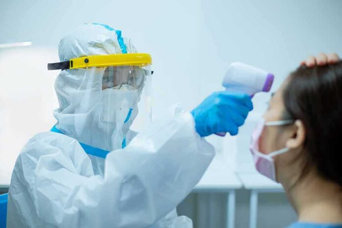 shutterstock_1689807307 Healthcare worker in PPE checks patient temperature with infrared thermometer