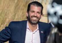 Donald Trump Jr. smiling in a navy suit outdoors