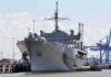 Large gray naval ship docked at an industrial port